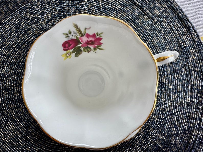 Puede incluir: Una taza de t&eacute; blanca con un borde dorado y un dise&ntilde;o floral con rosas rosas y rojas. La taza de t&eacute; tiene un borde festoneado y un asa.