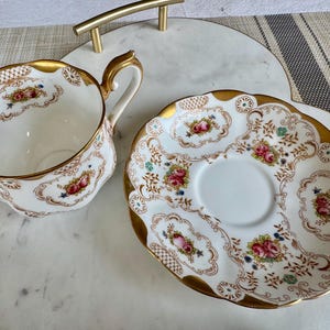 K&ouml;nnte beinhalten: Ein Vintage-Tee-Set. Die wei&szlig;e Porzellantasse und Untertasse haben einen goldenen Rand und ein Blumendesign mit rosa und roten Rosen. Das Set steht auf einem Marmortablett mit goldenem Griff.
