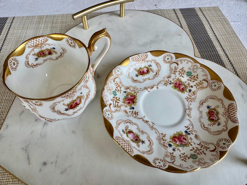 K&ouml;nnte beinhalten: Ein Vintage-Tee-Set. Die wei&szlig;e Porzellantasse und Untertasse haben einen goldenen Rand und ein Blumendesign mit rosa und roten Rosen. Das Set steht auf einem Marmortablett mit goldenem Griff.