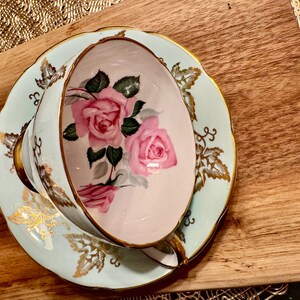 May include: A vintage teacup and saucer set with a delicate floral design. The cup and saucer are painted with pink roses and green leaves on a light blue background. The edges of the saucer are trimmed with gold.