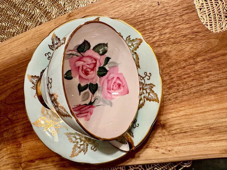 May include: A vintage teacup and saucer set with a delicate floral design. The cup and saucer are painted with pink roses and green leaves on a light blue background. The edges of the saucer are trimmed with gold.