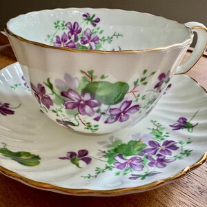 K&ouml;nnte beinhalten: Ein wei&szlig;es Tee-Set mit Blumendesign. Die Tasse und Untertasse sind mit lila Veilchen und gr&uuml;nen Bl&auml;ttern verziert. Die R&auml;nder sind goldfarben. Die Tasse hat einen Henkel.