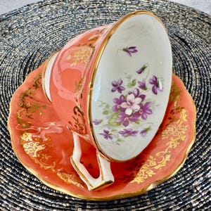May include: A coral pink teacup and saucer with a gold trim and floral design. The cup features a white interior with purple and white flowers.