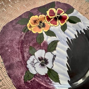 May include: A square decorative plate with a floral design. The plate features a dark purple rim and a white, blue, and black center. Three pansies in yellow, red, and white with green leaves are painted on the plate.