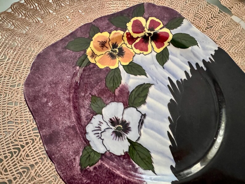 May include: A square decorative plate with a floral design. The plate features a dark purple rim and a white, blue, and black center. Three pansies in yellow, red, and white with green leaves are painted on the plate.
