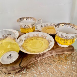 Puede incluir: Un juego de tazas y platillos vintage. Las tazas y los platillos son blancos con detalles florales dorados y una banda amarilla brillante. Las tazas tienen asas curvas. El juego est&aacute; dispuesto sobre una tabla de madera.