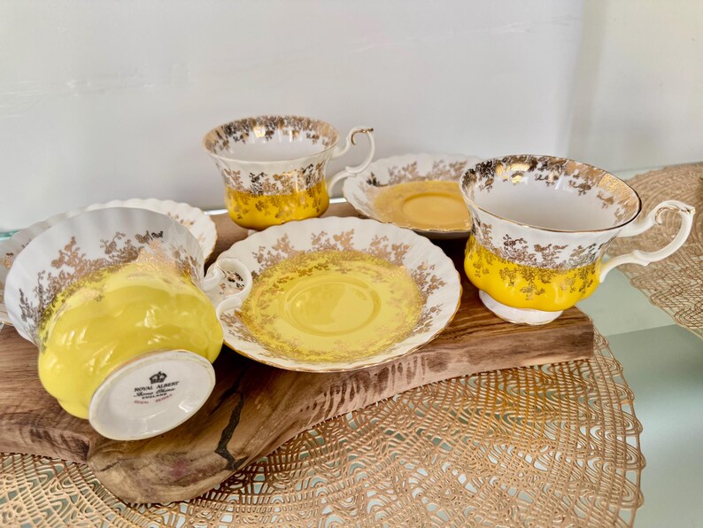 Puede incluir: Un juego de tazas y platillos vintage. Las tazas y los platillos son blancos con detalles florales dorados y una banda amarilla brillante. Las tazas tienen asas curvas. El juego est&aacute; dispuesto sobre una tabla de madera.