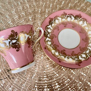 May include: A pink teacup and saucer set with gold filigree detailing. The cup has a curved handle and a gold rim. The saucer has a matching design. The set is on a textured, gold-colored surface. A classic tea set.
