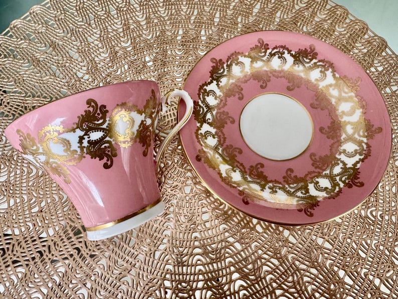 May include: A pink teacup and saucer set with gold filigree detailing. The cup has a curved handle and a gold rim. The saucer has a matching design. The set is on a textured, gold-colored surface. A classic tea set.