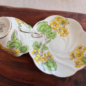 May include: A white teacup and saucer set with a floral design featuring yellow flowers and green leaves. The teacup has a gold rim and the saucer has a scalloped edge.