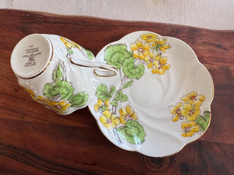 May include: A white teacup and saucer set with a floral design featuring yellow flowers and green leaves. The teacup has a gold rim and the saucer has a scalloped edge.