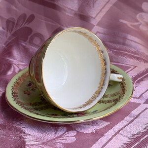 May include: A light green porcelain teacup and saucer set featuring a white interior, gold gilded rims, and a delicate floral pattern with gold leaf accents along the edges of both the cup and the saucer, showcasing a classic scalloped design.