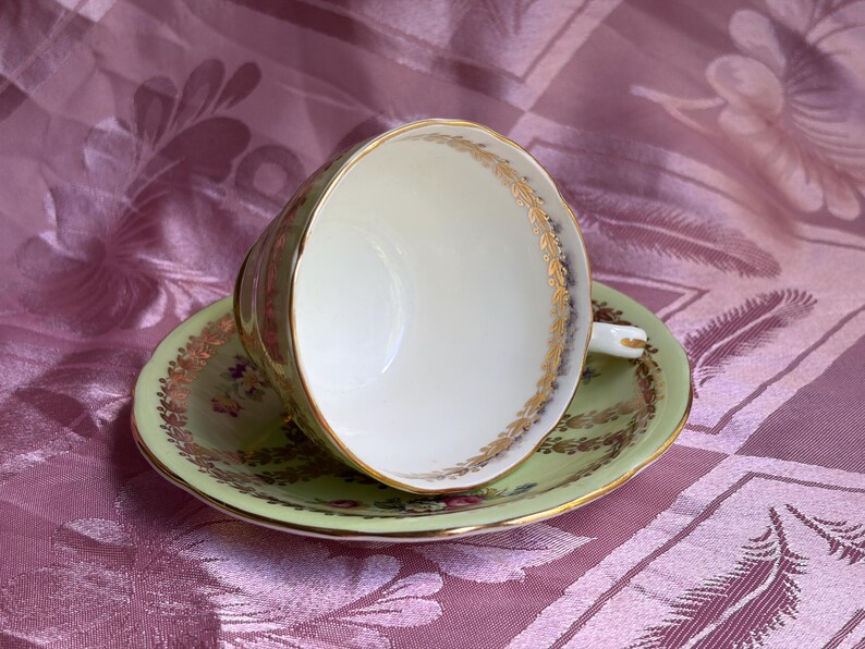 May include: A light green porcelain teacup and saucer set featuring a white interior, gold gilded rims, and a delicate floral pattern with gold leaf accents along the edges of both the cup and the saucer, showcasing a classic scalloped design.