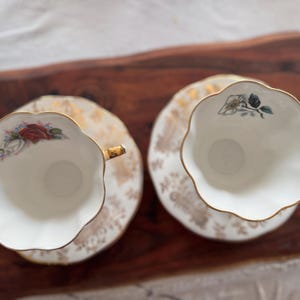 May include: Two white teacups with gold trim and floral designs on the side. The cups are on matching saucers with gold trim and floral designs.