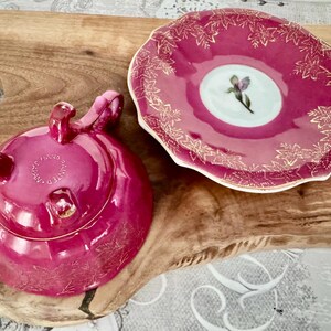 May include: A magenta-colored ceramic teapot with a matching saucer. The teapot has a lid and three small handles. The saucer has a floral design with gold accents. The teapot and saucer are on a wooden surface.