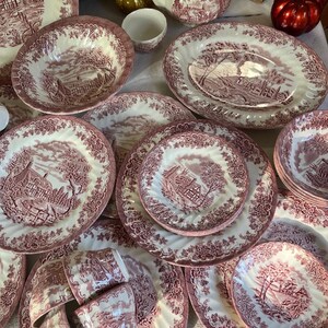 Pu&ograve; includere: Una collezione di stoviglie antiche bianche e bordeaux, tra cui piatti, ciotole, tazze e una teiera. Ogni pezzo presenta una scena paesaggistica dettagliata e un bordo floreale decorativo. Il set &egrave; disposto su una superficie bianca.