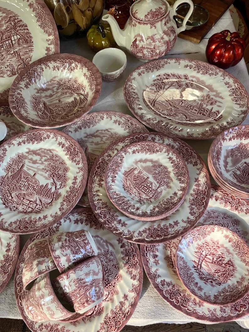 Pu&ograve; includere: Una collezione di stoviglie antiche bianche e bordeaux, tra cui piatti, ciotole, tazze e una teiera. Ogni pezzo presenta una scena paesaggistica dettagliata e un bordo floreale decorativo. Il set &egrave; disposto su una superficie bianca.