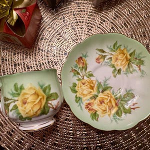 May include: A green teacup and saucer with a yellow rose floral pattern. The saucer has a scalloped edge.