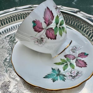May include: White teacup and saucer set with a floral design featuring pink and green leaves and white flowers. The cup and saucer have gold trim. The set is displayed on a silver serving tray.