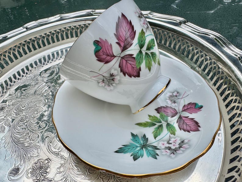 May include: White teacup and saucer set with a floral design featuring pink and green leaves and white flowers. The cup and saucer have gold trim. The set is displayed on a silver serving tray.