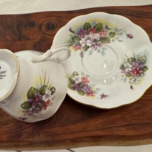 May include: A white teacup and saucer set with a floral pattern of purple and white violets. The saucer has a scalloped edge and the teacup has a handle. The set is decorated with gold trim. The teacup has a mark on the bottom that reads "Royal Albert Bone China England".