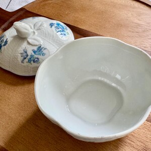 May include: A white ceramic bowl with a scalloped edge and a matching lid. The lid features a floral design in blue and white. The bowl and lid are on a wooden tray.