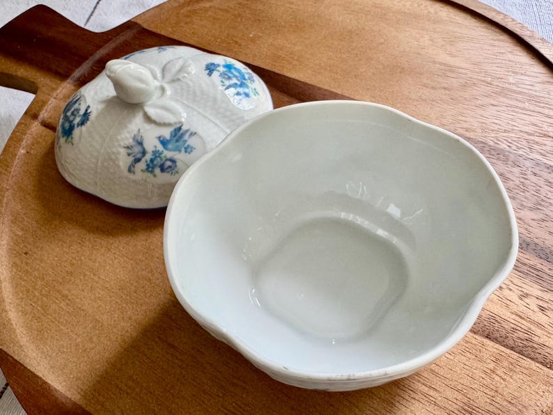 May include: A white ceramic bowl with a scalloped edge and a matching lid. The lid features a floral design in blue and white. The bowl and lid are on a wooden tray.
