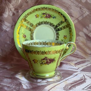 May include: A light green teacup and matching saucer set featuring gold-toned leaf patterns and floral motifs. The teacup has a scalloped rim and a curved handle, while the saucer displays a circular arrangement of gold leaves and small flowers.