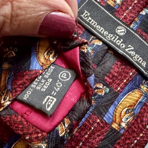 May include: An Ermenegildo Zegna necktie featuring a patterned design of gold, blue, and maroon geometric shapes. The tie includes a black brand label and a care tag indicating 100% silk, with a vibrant magenta fabric lining visible at the center.
