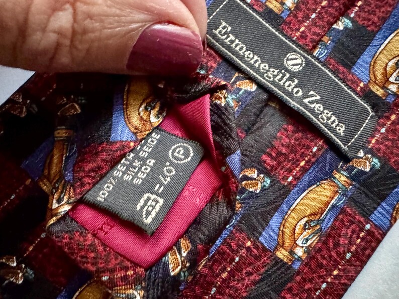 May include: An Ermenegildo Zegna necktie featuring a patterned design of gold, blue, and maroon geometric shapes. The tie includes a black brand label and a care tag indicating 100% silk, with a vibrant magenta fabric lining visible at the center.