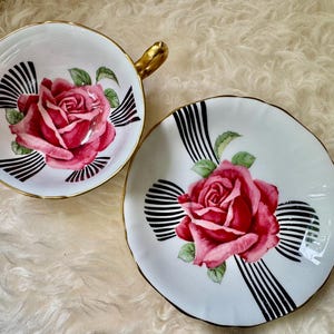May include: A white teacup and saucer set with a pink rose design. The teacup has a gold handle and trim, and the saucer has a matching design. Black and white striped accents surround the rose. A classic tea set.