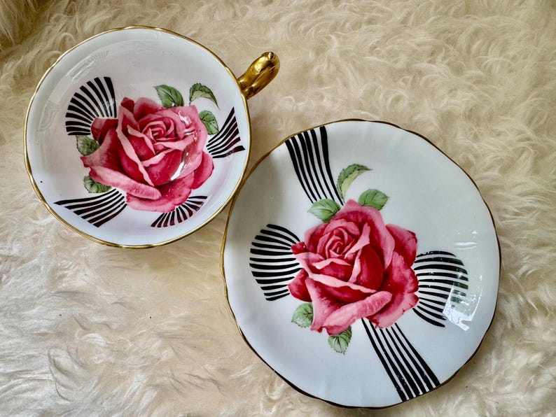 May include: A white teacup and saucer set with a pink rose design. The teacup has a gold handle and trim, and the saucer has a matching design. Black and white striped accents surround the rose. A classic tea set.