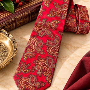 May include: A vibrant red necktie featuring an intricate, multi-colored paisley pattern in shades of gold, dark brown, and muted blue. The tie has a pointed tip and a smooth, satin-like finish, displaying a repeating teardrop-shaped motif throughout.