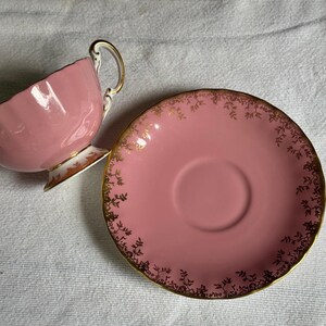 Peut inclure: Un service &agrave; th&eacute; rose compos&eacute; d'une tasse et d'une soucoupe. La tasse est inclin&eacute;e, r&eacute;v&eacute;lant son int&eacute;rieur. La soucoupe est ronde, rose et orn&eacute;e d'un motif floral dor&eacute; sur le bord. La poign&eacute;e et la base de la tasse sont &eacute;galement dor&eacute;es.