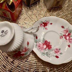 May include: A white teacup and saucer with a floral pattern of pink and red roses. The teacup has the words "Royal Albert Bone China England" on the bottom.