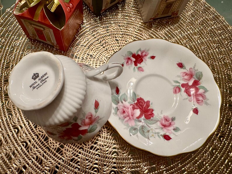 May include: A white teacup and saucer with a floral pattern of pink and red roses. The teacup has the words "Royal Albert Bone China England" on the bottom.