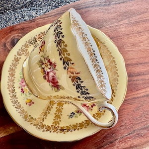 May include: A pale yellow porcelain teacup and saucer set featuring a delicate floral pattern with pink and yellow roses. The rim and handle are accented with intricate gold leaf filigree detailing, creating a refined and elegant appearance.