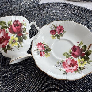 Puede incluir: Un juego de taza y platillo de t&eacute; blanco con un patr&oacute;n floral con rosas rojas, rosas y amarillas. El juego tiene un borde dorado.