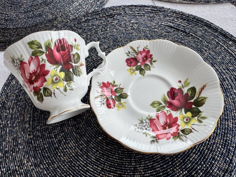 Puede incluir: Un juego de taza y platillo de t&eacute; blanco con un patr&oacute;n floral con rosas rojas, rosas y amarillas. El juego tiene un borde dorado.