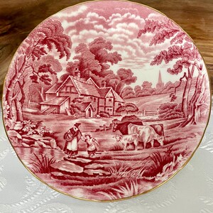 Pu&ograve; includere: Un piatto rotondo rosso e bianco con una scena pastorale. Il design presenta una fattoria, alberi, figure e bestiame. Il piatto ha un bordo dorato ed &egrave; posto su uno sfondo bianco.