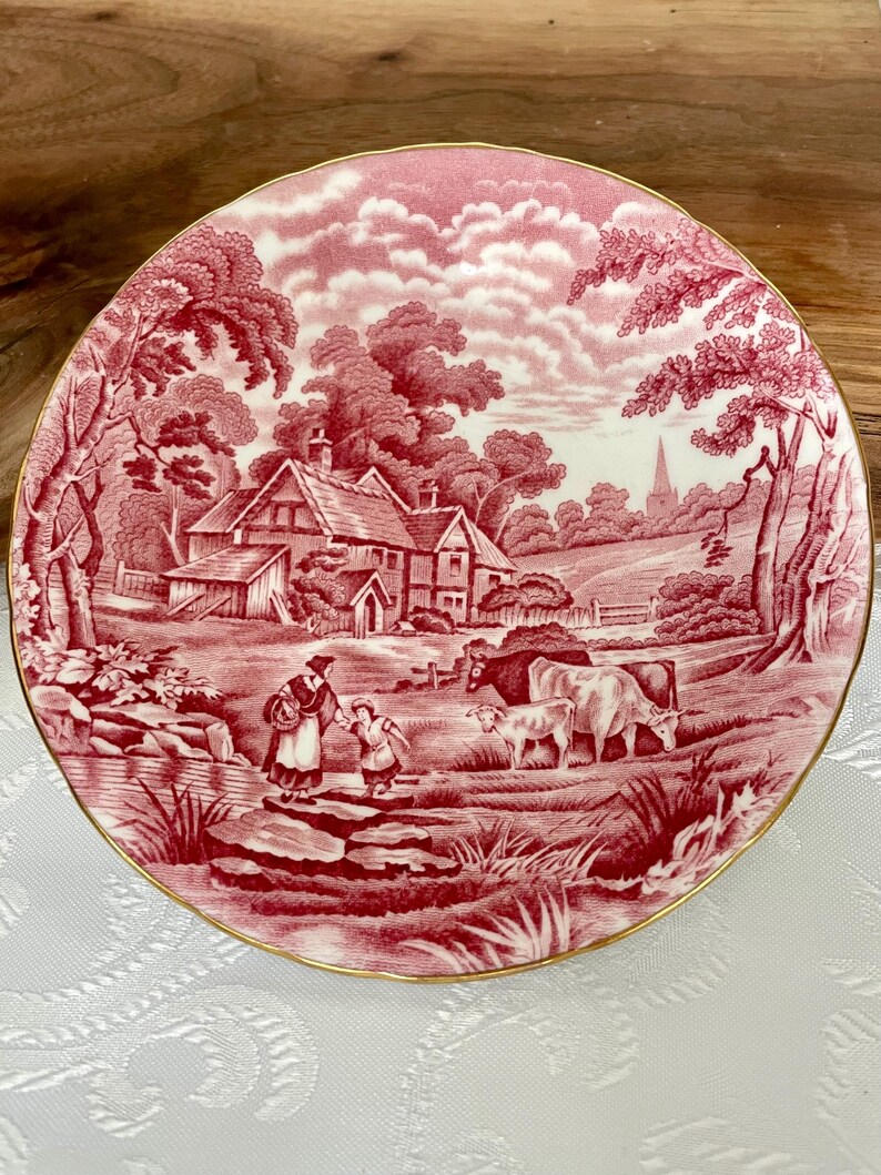 Pu&ograve; includere: Un piatto rotondo rosso e bianco con una scena pastorale. Il design presenta una fattoria, alberi, figure e bestiame. Il piatto ha un bordo dorato ed &egrave; posto su uno sfondo bianco.