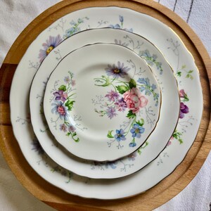 May include: Three white porcelain plates with a floral pattern of pink roses, purple and blue flowers, and gold edging. The plates are displayed on a round wooden serving tray.