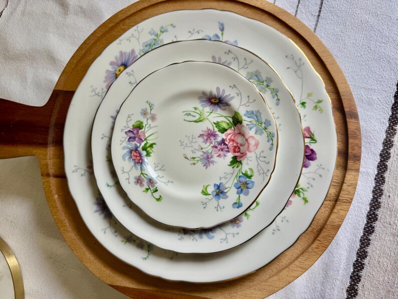 May include: Three white porcelain plates with a floral pattern of pink roses, purple and blue flowers, and gold edging. The plates are displayed on a round wooden serving tray.