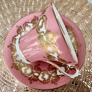May include: A pink teacup and saucer set with gold filigree detailing. The teacup is tilted, revealing a white interior and a gold-trimmed rim. The saucer mirrors the cup's design, creating a cohesive and elegant aesthetic.