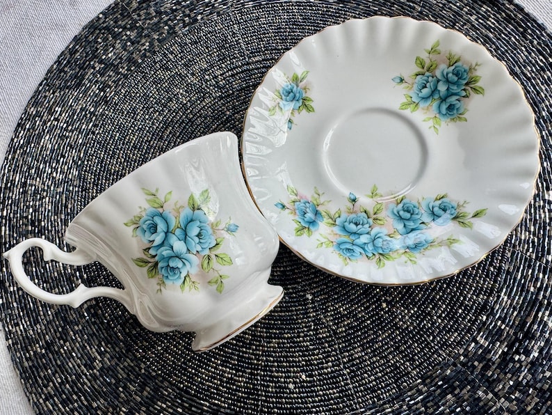 May include: A white teacup and saucer set with a blue floral pattern. The teacup has a scalloped rim and a delicate handle. The saucer has a scalloped edge and a floral design around the rim.
