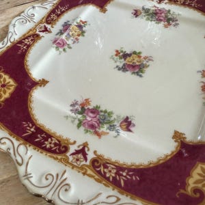 May include: A white square serving tray with a burgundy border and gold accents. The tray is decorated with a floral pattern in shades of pink, yellow, and green.