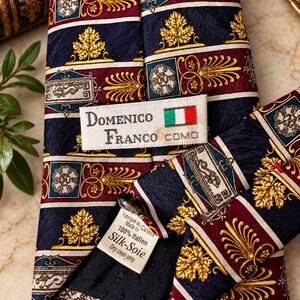 May include: A navy blue silk necktie featuring a repeating pattern of gold leaves, burgundy stripes, and rectangular motifs with floral designs. A white label reads Domenico Franco Como with an Italian flag icon and a tag stating 100% Silk.