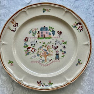 May include: Cream-colored ceramic plate with a brown rim and scalloped edges. The center features a farm scene with a pink house, animals, and a tree. Floral and animal motifs are scattered around the plate's edge.