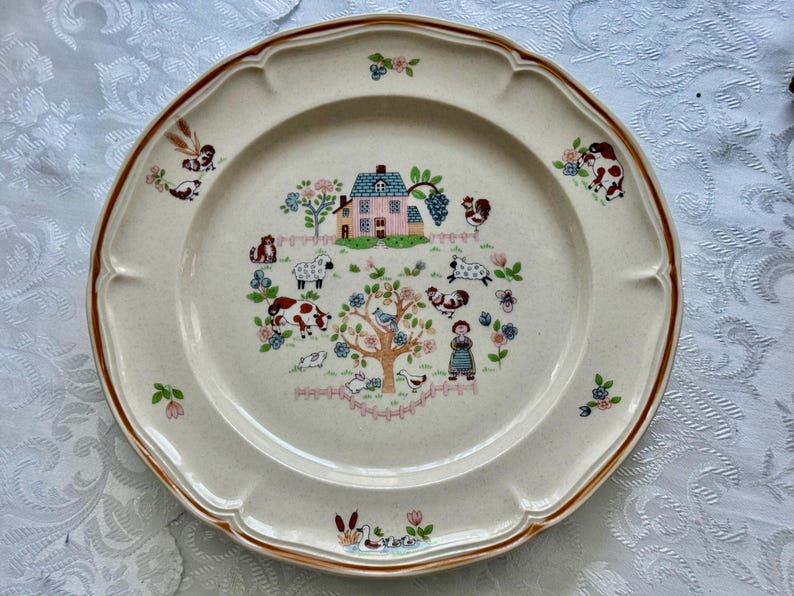 May include: Cream-colored ceramic plate with a brown rim and scalloped edges. The center features a farm scene with a pink house, animals, and a tree. Floral and animal motifs are scattered around the plate's edge.