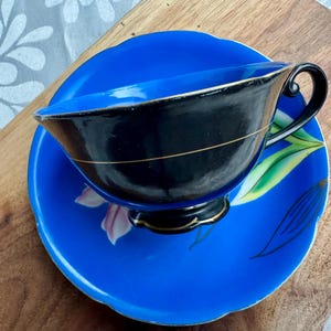 Puede incluir: Un juego de taza y platillo de t&eacute; azul y negro con un dise&ntilde;o floral. La taza de t&eacute; tiene un interior negro y un borde dorado. El platillo tiene un fondo azul con un dise&ntilde;o floral rosa y verde.